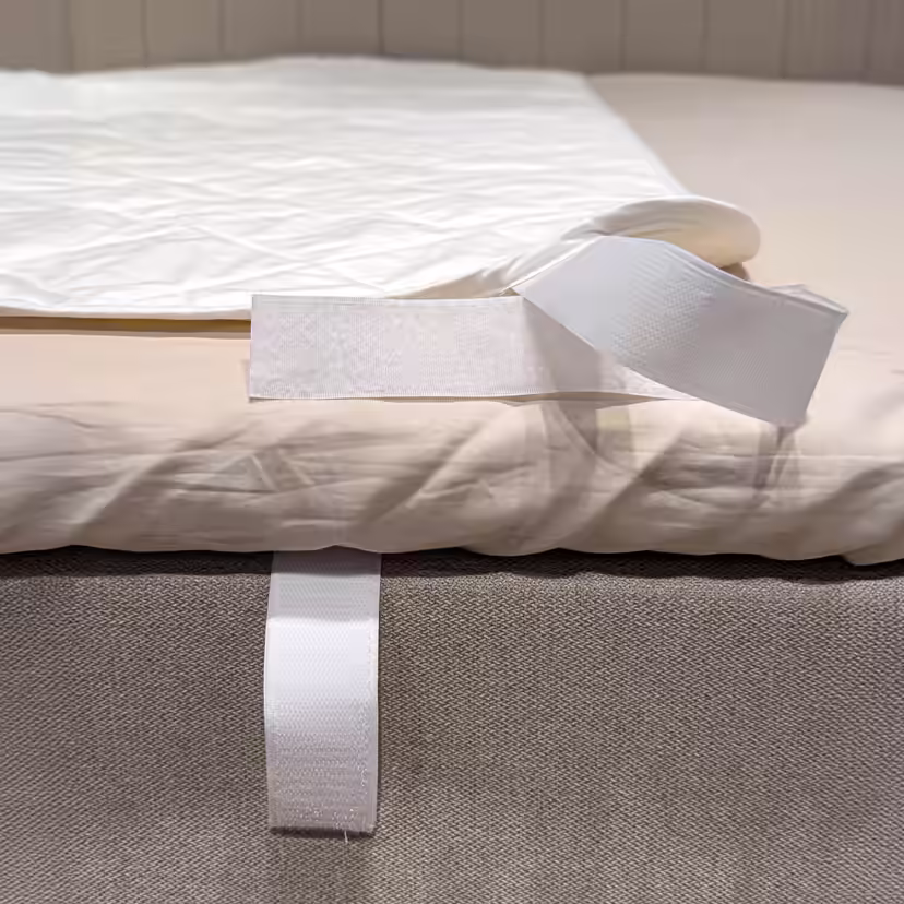 Corner of the white quilted mattress protector folded back to reveal the hook-and-loop strap around the edge of the bed
